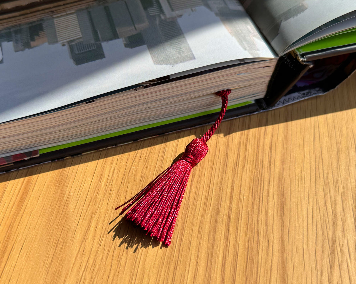 Bookmark tucked inside a closed hardcover book, tassel visible at the top. Thin aluminum design protects pages while adding style. Thoughtful gift for book lovers, readers, students, and anyone who enjoys unique and artistic bookmarks.