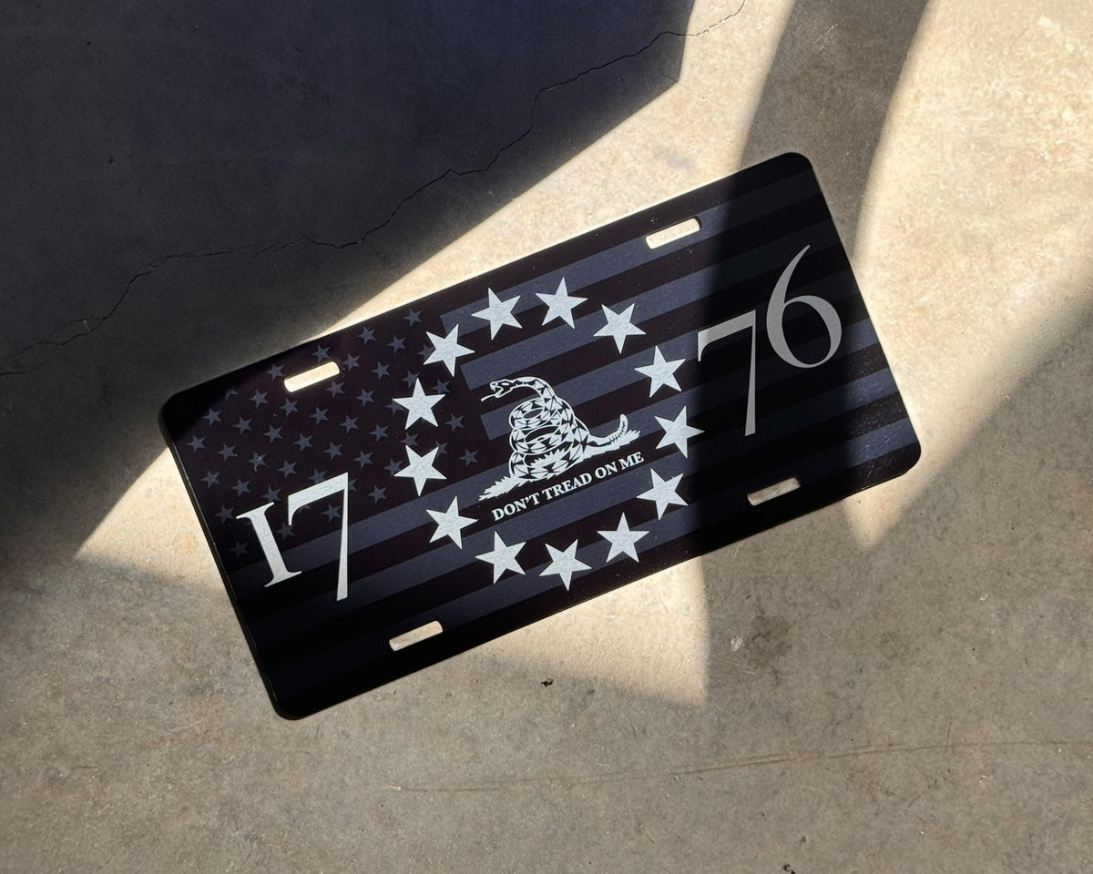 A black novelty license plate featuring a distressed American flag design with white stars, a Gadsden flag motif, and the numbers 1776, photographed on a concrete surface in direct sunlight.