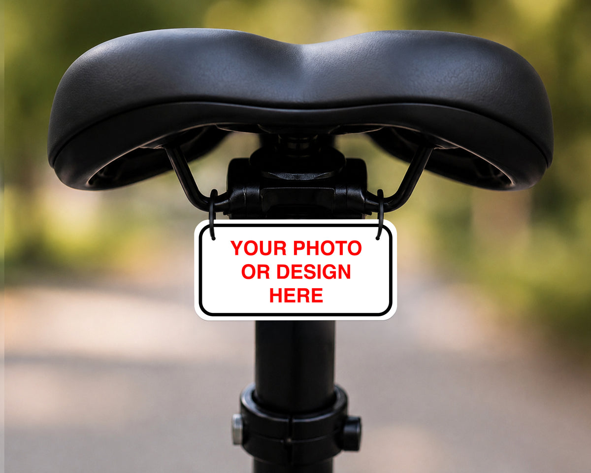 A customizable black bicycle saddle with a central mounting bracket holding a white license plate sign that reads 'YOUR PHOTO OR DESIGN HERE' in red text, set against a blurred outdoor background.