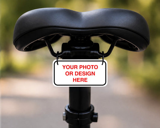 A customizable black bicycle saddle with a central mounting bracket holding a white license plate sign that reads 'YOUR PHOTO OR DESIGN HERE' in red text, set against a blurred outdoor background.