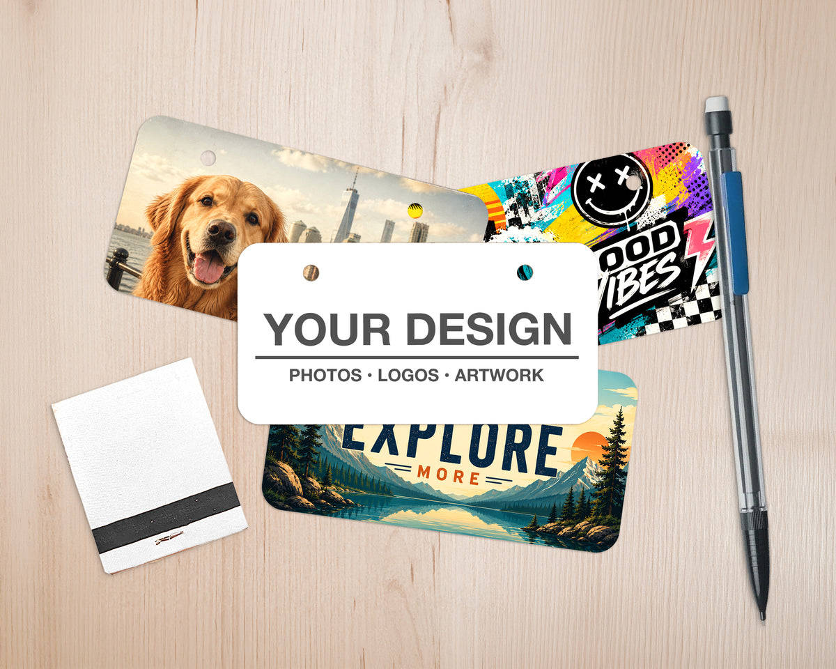 A styled flat-lay mockup on a light wood surface showing a customizable mini bicyclelicense plate with a golden retriever, a colorful graphic ID card, a scenic landscape ID card, a white notepad with a black stripe, and a pen.