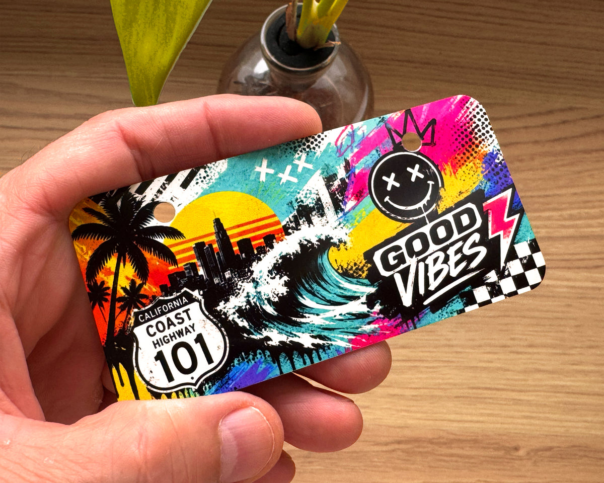 A hand holding a colorful rectangular 4x2 inch mini license plate  featuring a retro California Coast Highway 101 theme with palm trees, a wave, a smiley face, and the text 'GOOD VIBES' and 'COAST HIGHWAY 101', shown over a wooden tabletop with a small vase in the background.