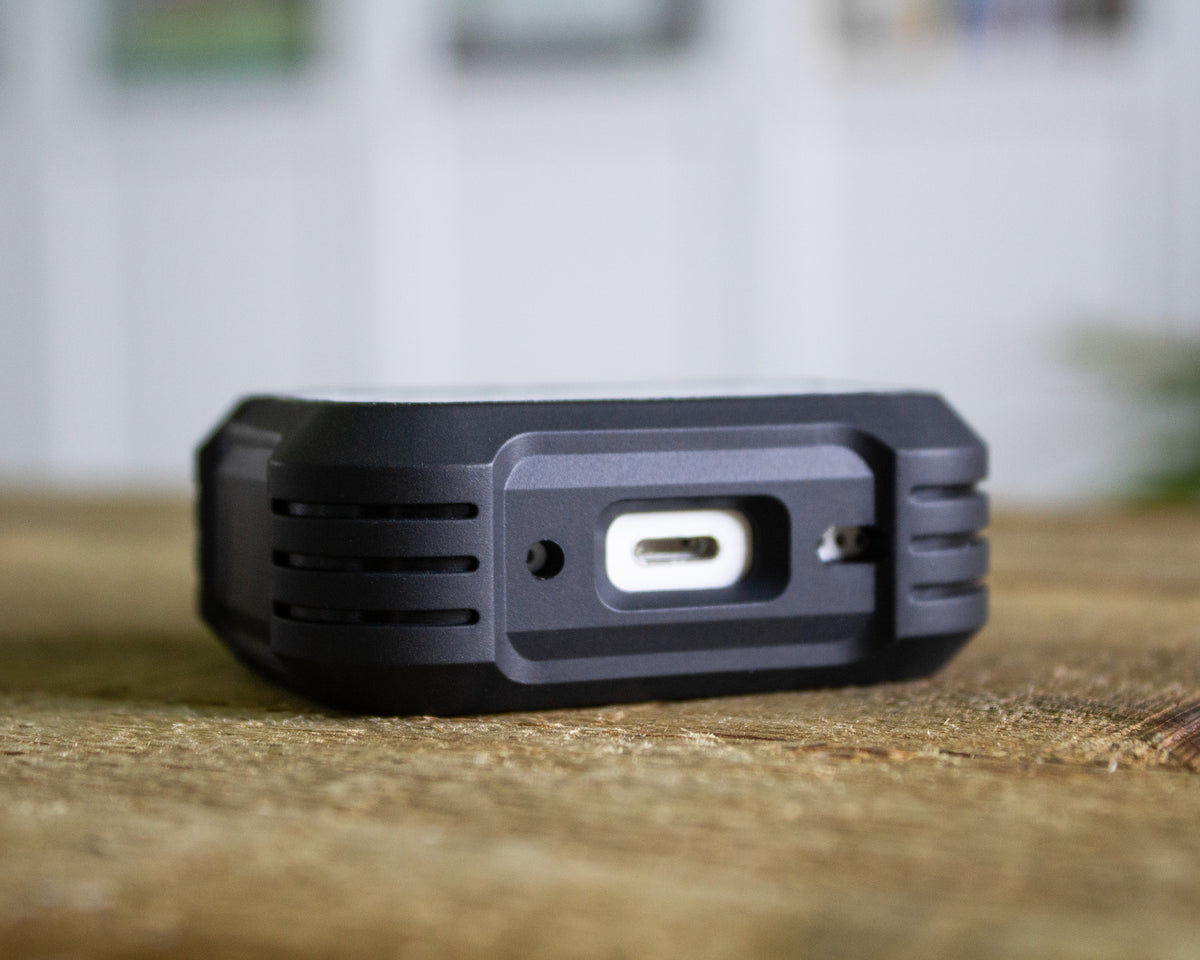 A close-up shot of a black rugged AirPods Pro 1st 2nd or 3rd Gen case, showing a USB-C port on its side, resting on a wooden surface with a blurred indoor background.