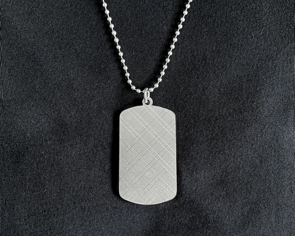 A silver dog tag pendant necklace with a textured grid pattern, hanging from a silver ball chain against a black fabric background.