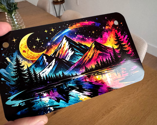 A rectangular metal license plate with rounded corners is held by hands over a wooden table. The plate features a highly detailed, colorful psychedelic landscape illustration with mountains, a lake, pine trees, a crescent moon, and stars in a galaxy theme.