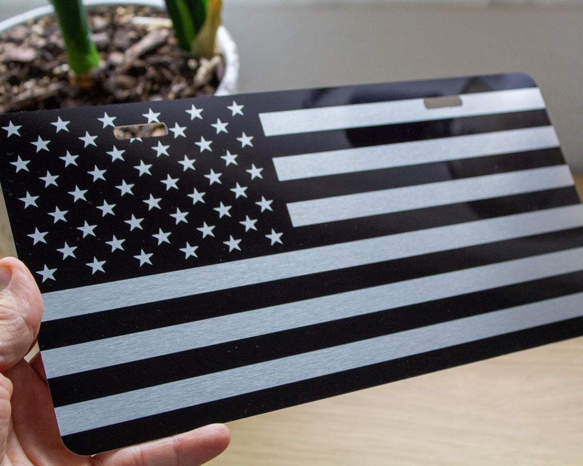A hand holds a black metal license plate featuring a brushed silver American flag design, with a blurred indoor background and a potted plant.