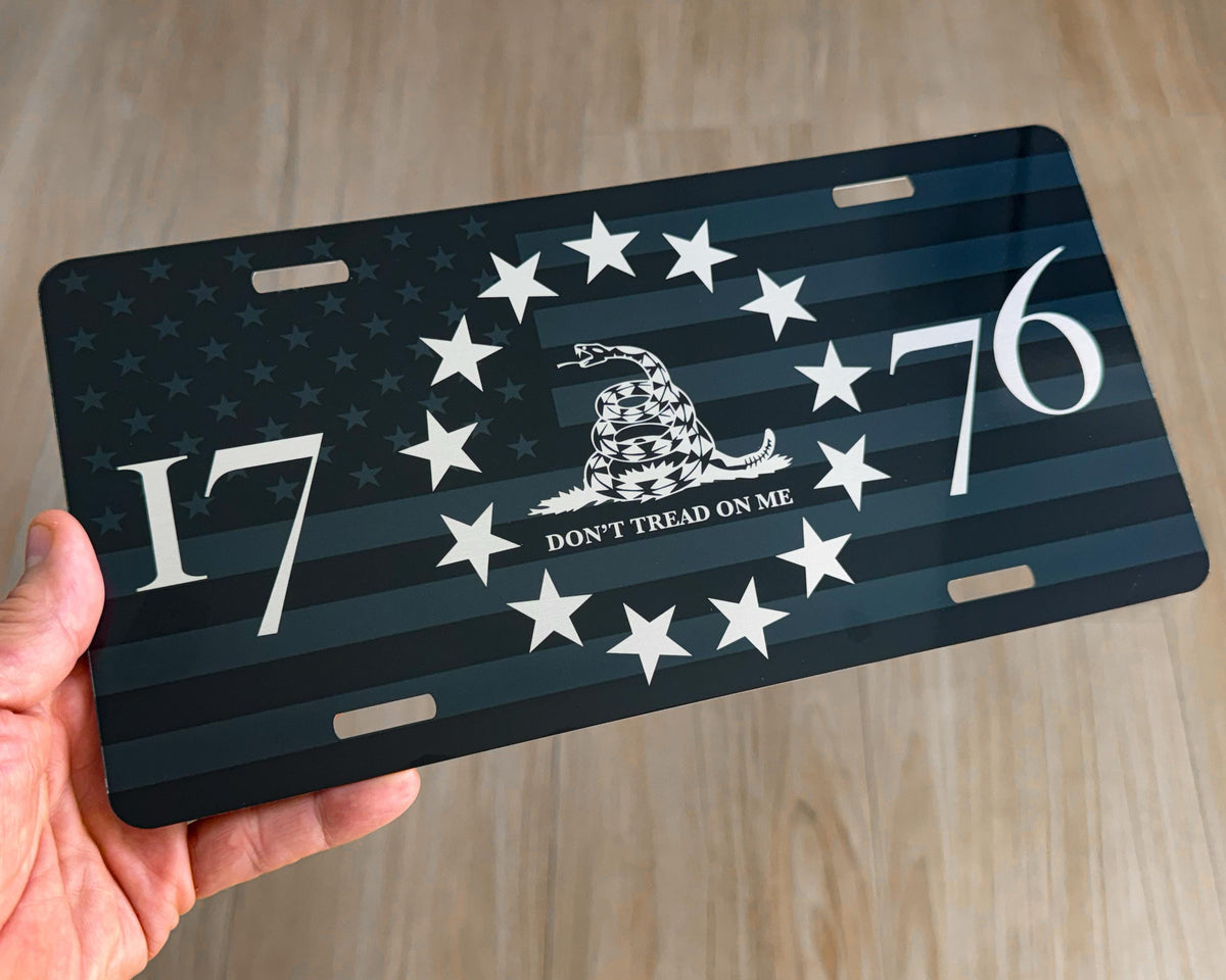 A hand holding a rectangular license plate featuring a dark blue and black American flag design with white stars, the text 'DON'T TREAD ON ME', the numbers 1776, and a coiled rattlesnake emblem in the center, against a light wood floor background.