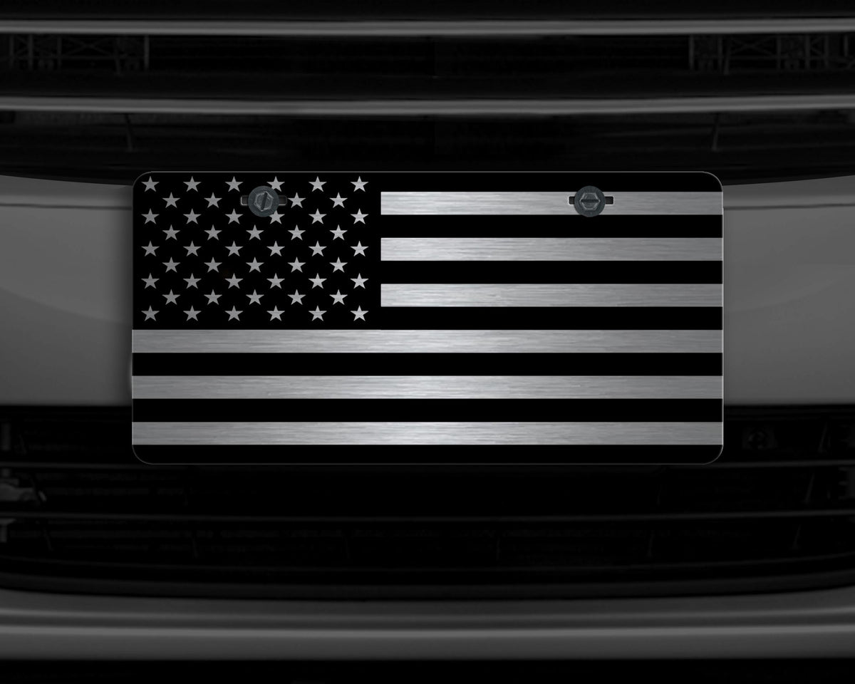 A license plate featuring a black and silver American flag design, mounted on a dark car grille.