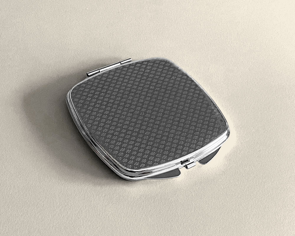 A silver-framed compact mirror showing a  textured pattern on the bottom on a neutral colored background