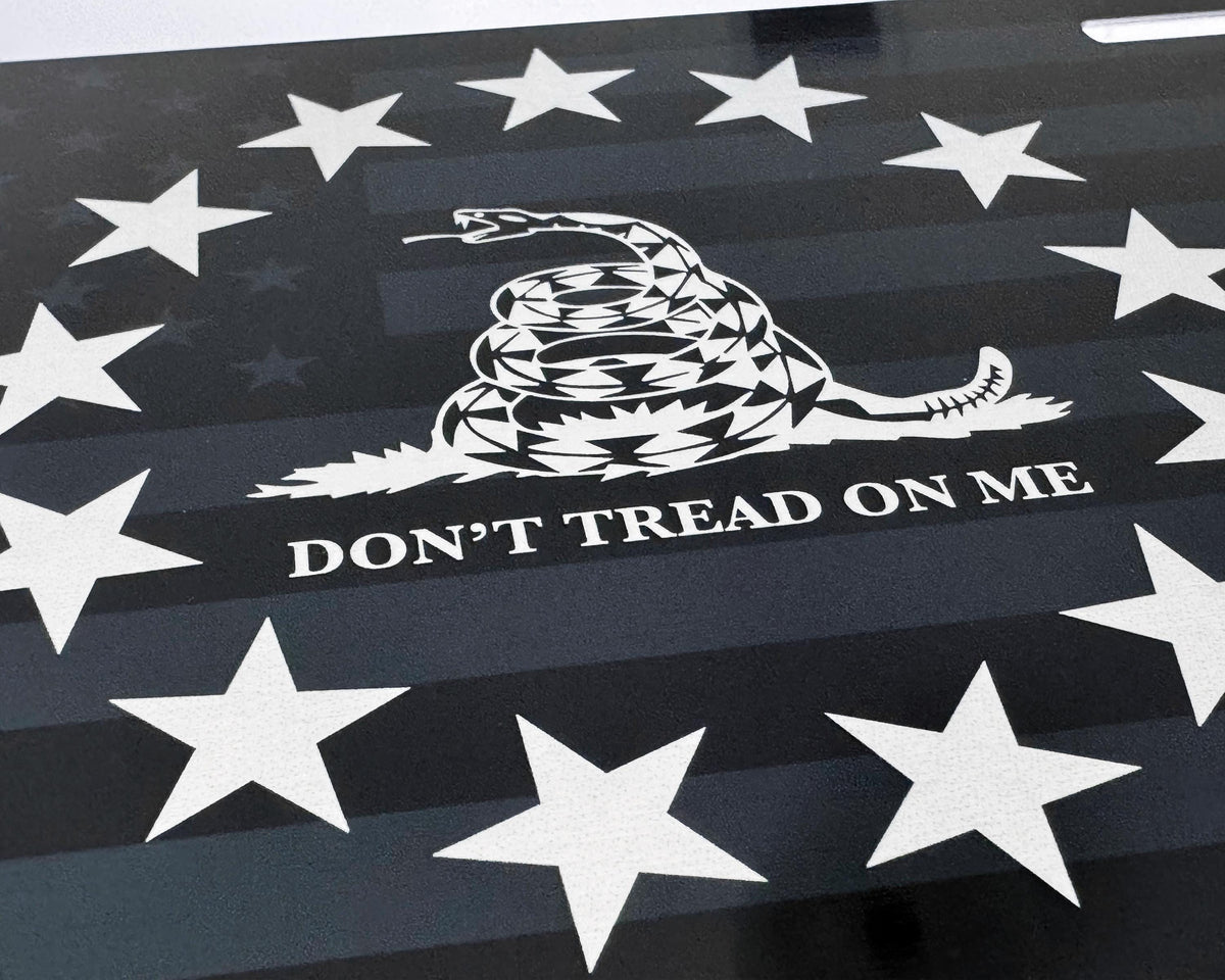 A close-up of a novelty license plate Gadsden flag design with a coiled rattlesnake, the text 'DON'T TREAD ON ME,' and multiple white stars on a silver black striped background.