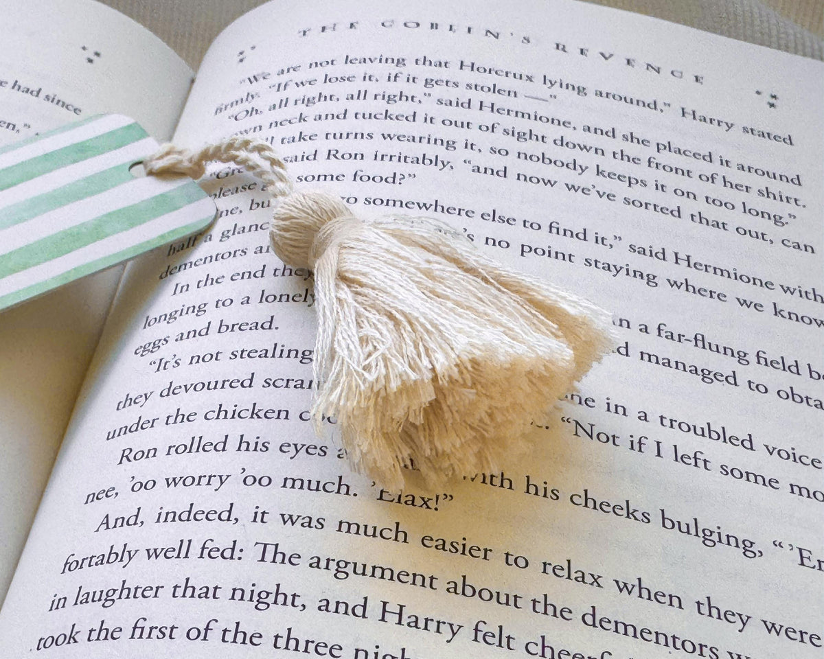 A cream-colored tassel bookmark made from cotton on an open book page, next to a striped paper bookmark