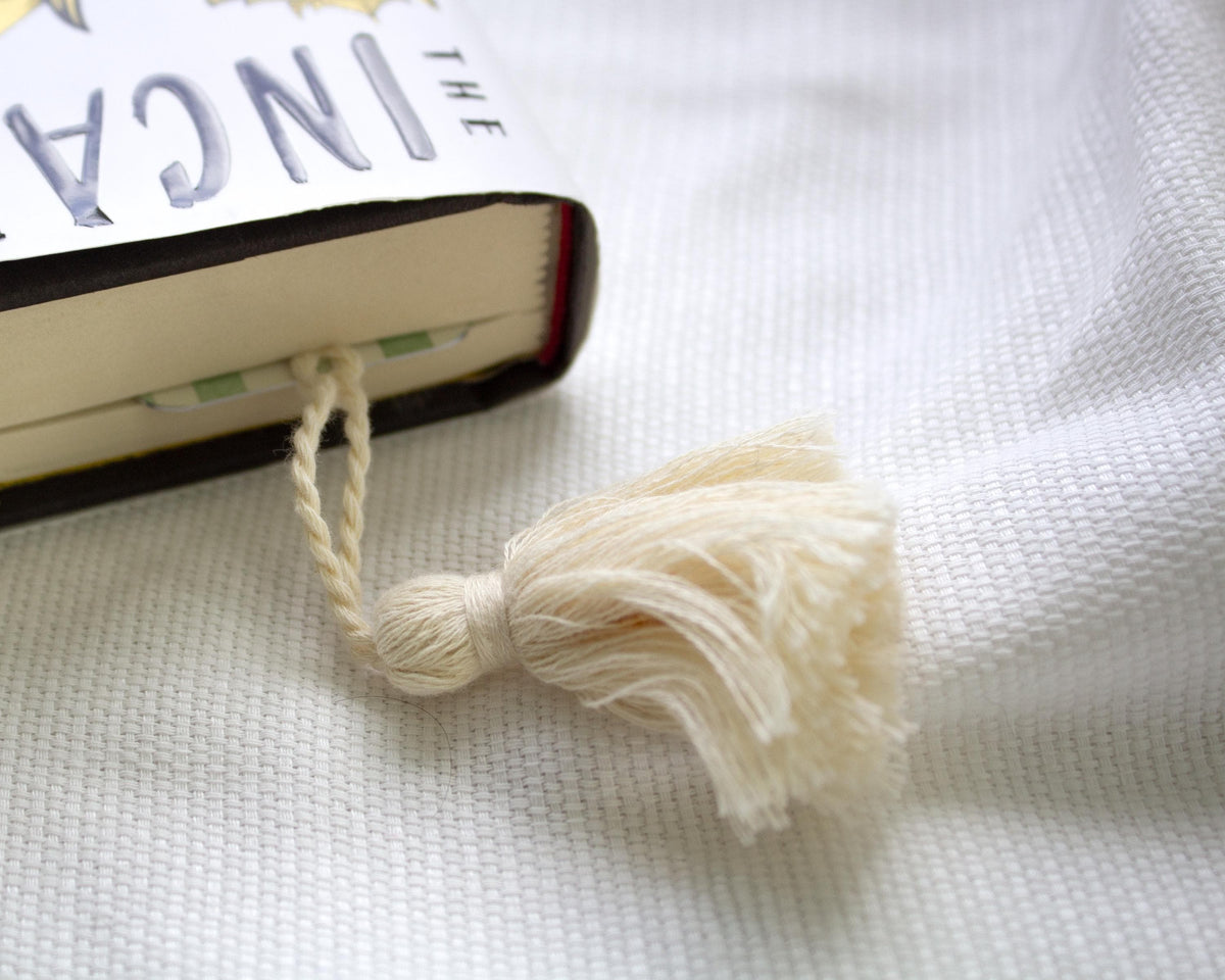 A cream-colored cotton tassel bookmark threaded through the page of a book on a textured white fabric background.