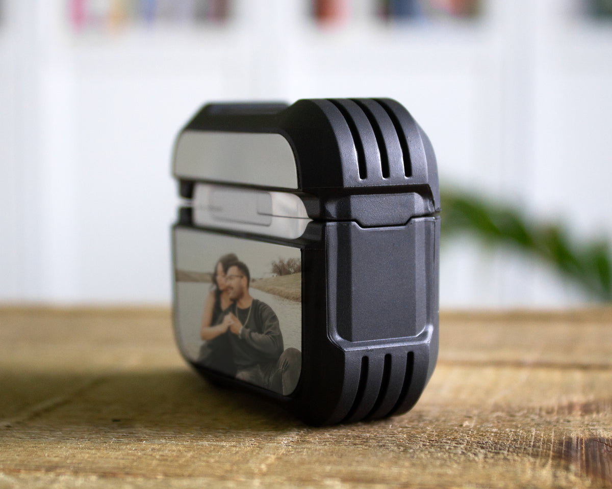A rugged black rubber AirPods Pro 1st 2nd or 3rd Gen case with a clear front window displaying a photo of a couple, sitting on a wooden tabletop with a blurred indoor background.