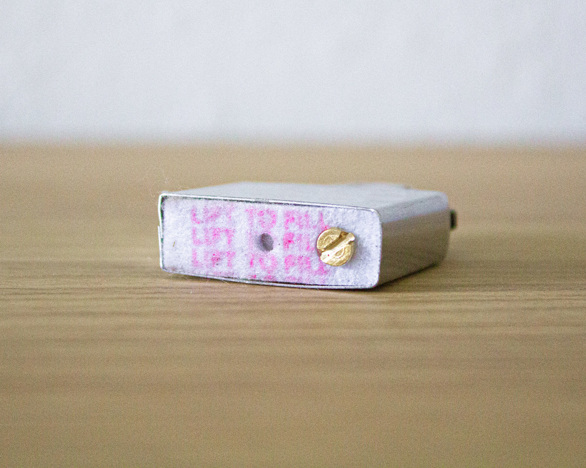 A refillable lighter insert with a pink patterned label and a gold screw, photographed on a light wood surface against a soft gray background.