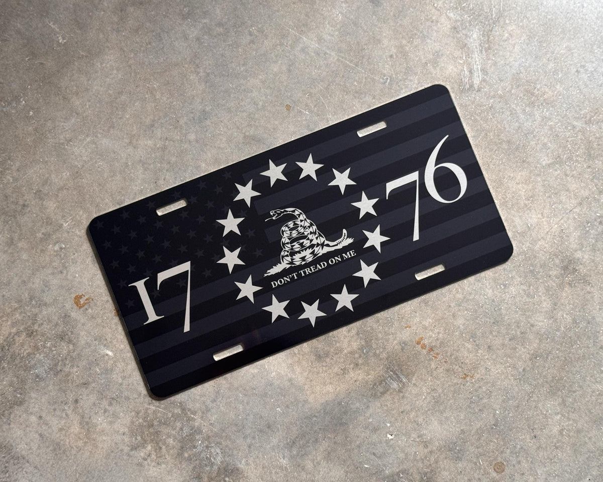 A black license plate featuring a distressed American flag design, a Gadsden flag motif with stars, and the numbers 1776, placed on a textured gray concrete surface.
