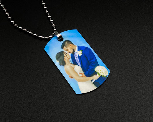 A custom dog tag necklace featuring a printed photo of a bride and groom kissing under a blue sky, hanging from a silver ball chain on a black background.