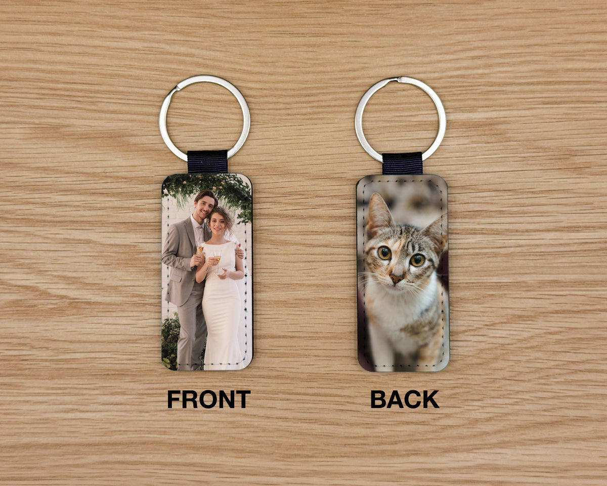 Two rectangular leather keychains shown on a wood surface, labeled FRONT and BACK. The front keychain displays a printed photo of a couple, and the back keychain displays a printed photo of a cat.