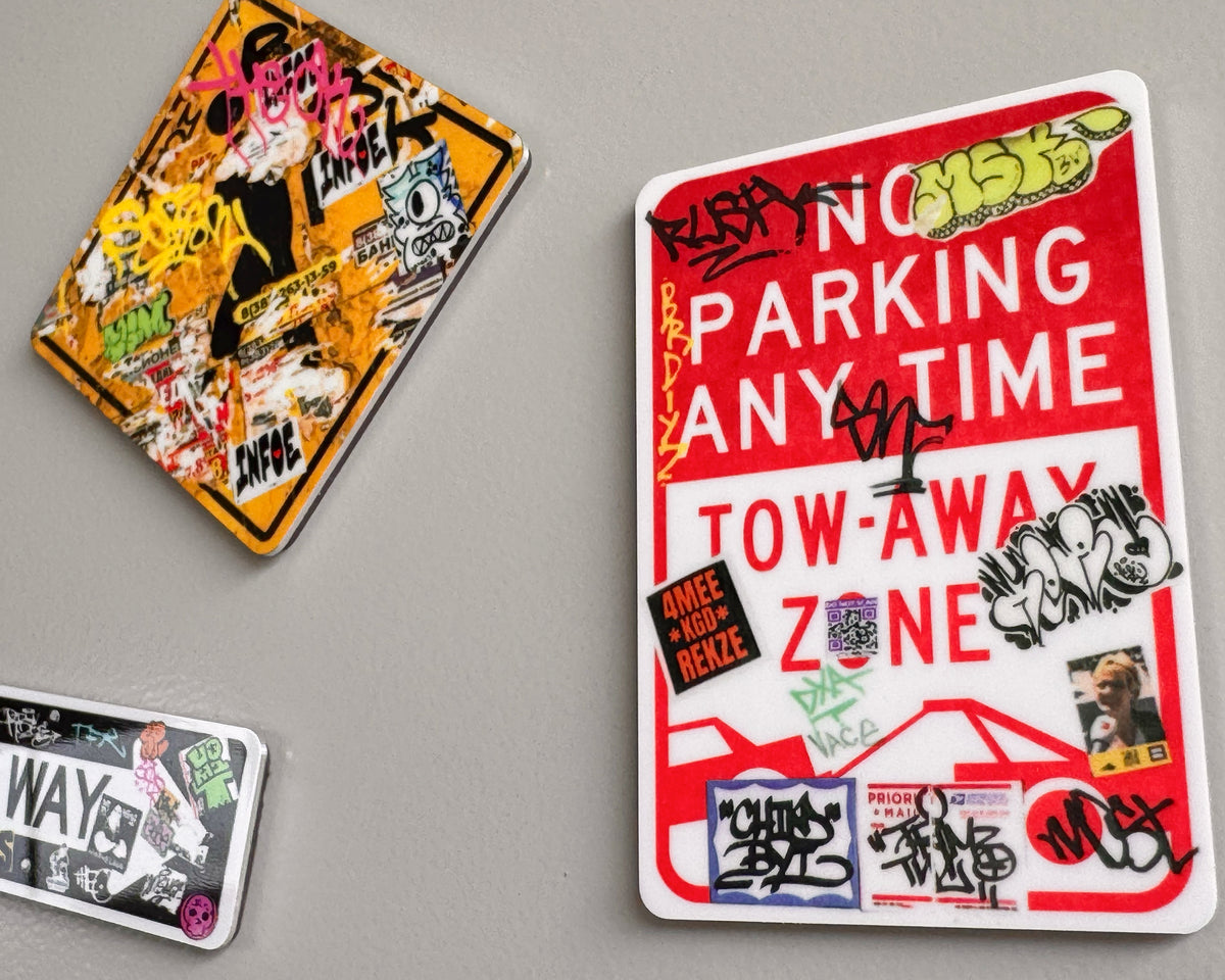 Three rectangular fridge magnets with graffiti-style designs and text, including a large red-and-white 'TOW-AWAY ZONE' sticker, displayed on a gray background.