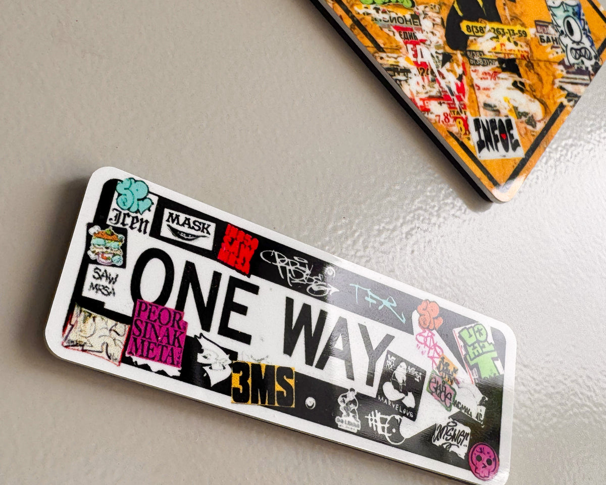 A rectangular white fridge magnet  graffiti covered with the words “ONE WAY” in large black letters, covered with many colorful graffiti-style stickers and patches, photographed on a light gray textured surface. Another similar sticker is partially visible at the top right.