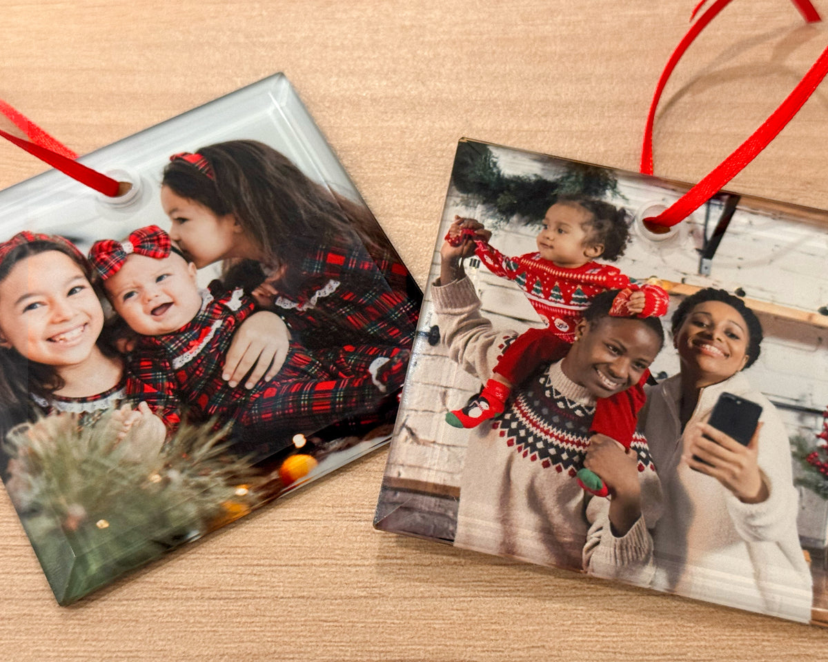 Two square glass photo ornaments with red hanging ribbons, displayed on a light wood surface. Each ornament features a printed family holiday photo, with one ornament showing a Christmas tree and the other a snowy outdoor scene.