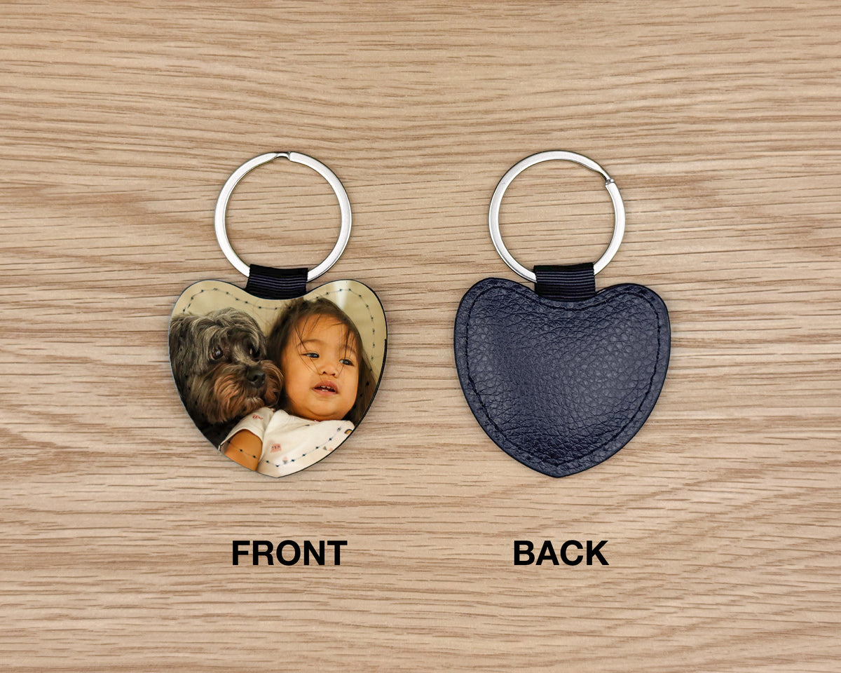 Two heart-shaped keychains shown on a light wood background, labeled FRONT and BACK. The left keychain shows a printed photo insert with a silver key ring and black tab; the right keychain shows the back with a dark navy textured surface and silver key ring.