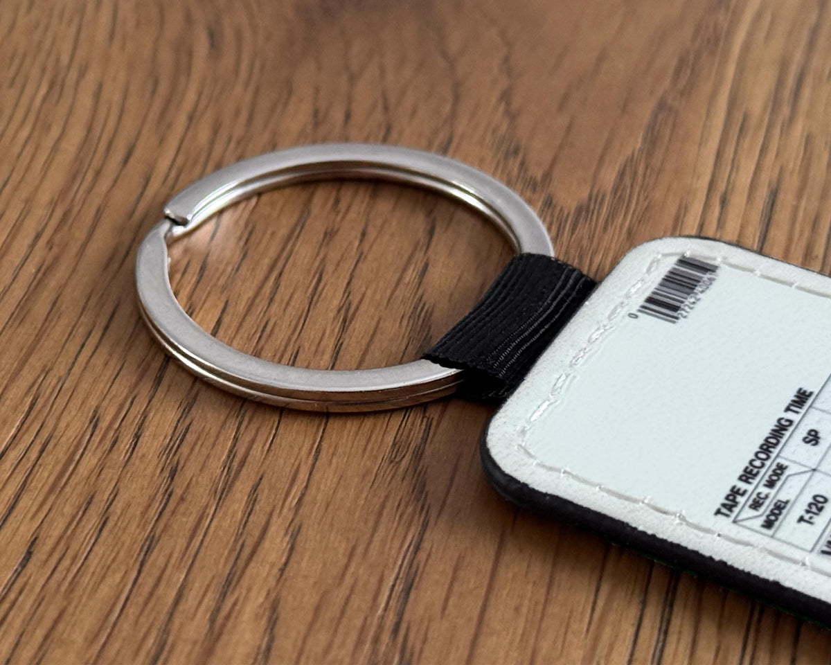 A close-up of a silver metal key ring attached to a black strap and a white rectangular VHS cassette keychain with printed text, placed on a brown wood-grain surface.