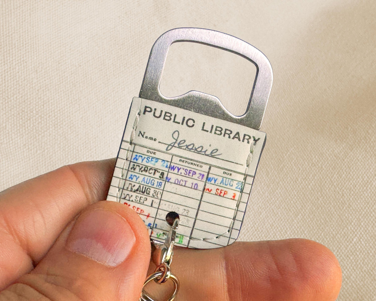 A novelty public library card keychain held in a hand. The keychain features a silver metal bottle opener at the top, a white card printed with 'PUBLIC LIBRARY' and a handwritten-style form, and a small metal keyring at the bottom.