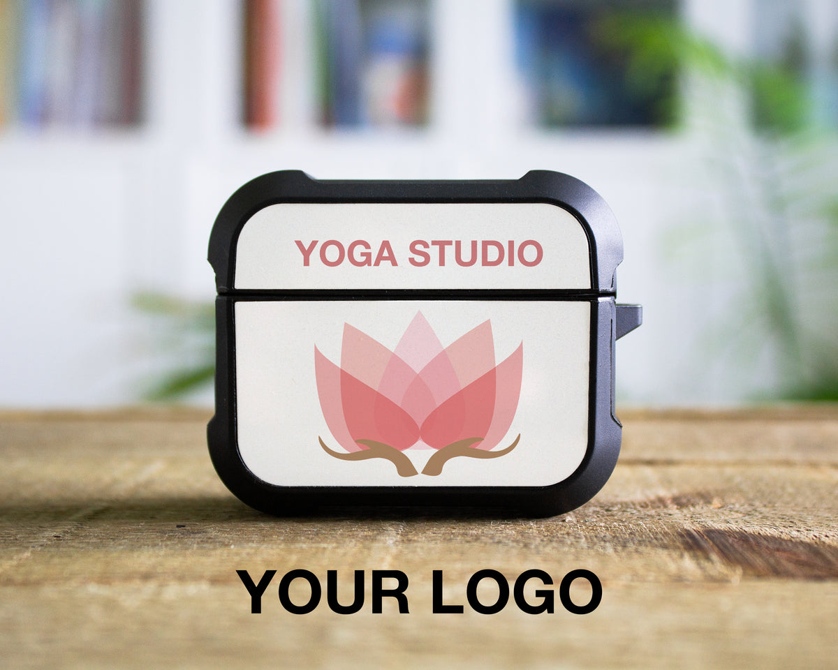 A custom protective AirPods Pro 1st 2nd or 3rd Gen case with a black bumper and a white back panel featuring a pink lotus flower graphic and the text 'YOGA STUDIO'. The case is shown standing on a wooden surface with a blurred indoor background, and the text 'YOUR LOGO' is printed below.