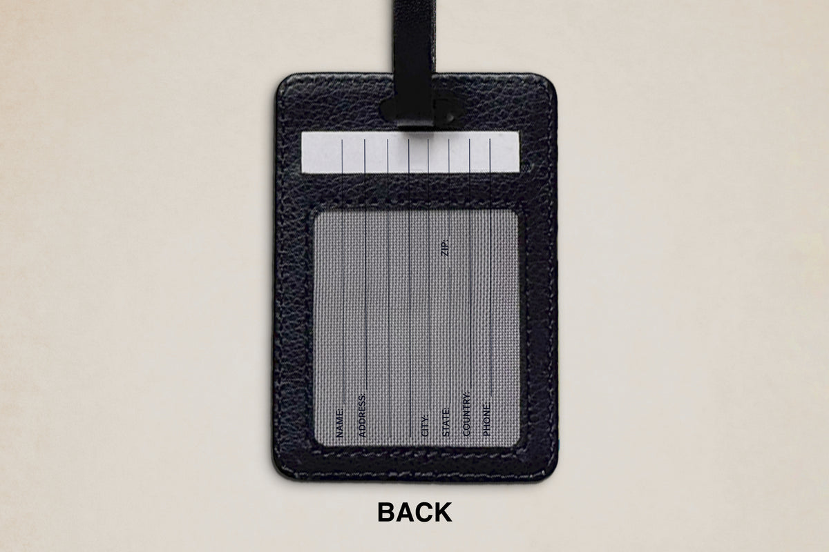 A black textured leather luggage tag shown from the back, featuring a black strap, a white address label area, and a gray transparent ID window with printed fields. The word 'BACK' is printed below the tag on a light beige background.