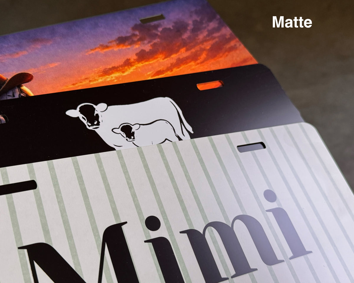 A close-up of a custom metal license plate with a matte finish, featuring a black horizontal stripe with a white cow and calf silhouette, a sunset sky graphic, and the name 'Mimi' in large black letters on a white and pale green striped background.