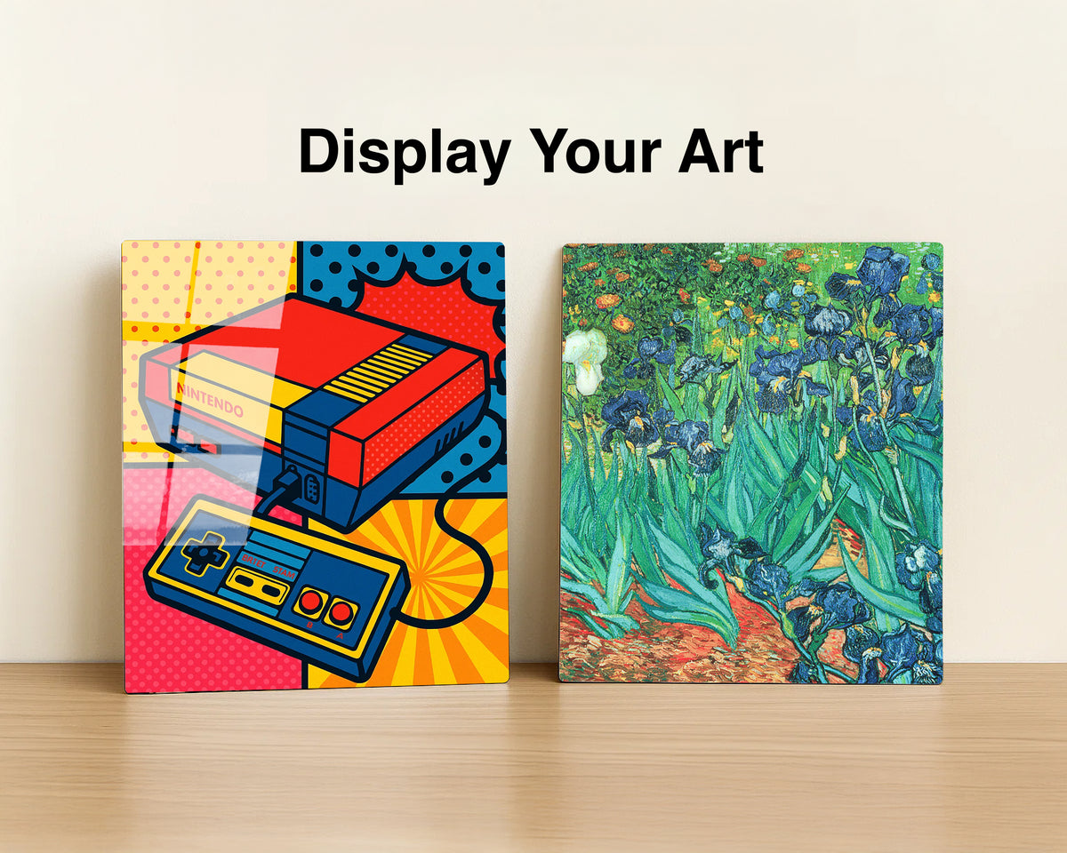 Two square art prints displayed side by side on a light wood surface against an off-white wall, with the text 'Display Your Art' above. The left print features a colorful pop-art retro game console and controller design.