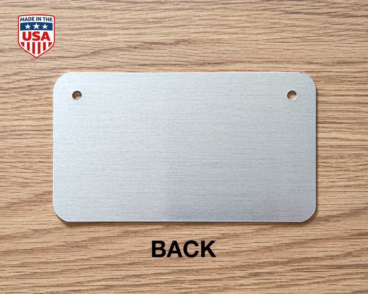 A brushed metal license plate blank with two pre-drilled holes, shown on a wood-grain background with the word 'BACK' below it and a 'Made in the USA' badge in the top-left corner.