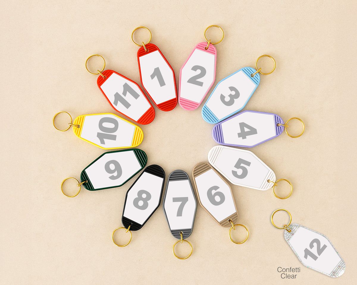 Twelve numbered keychain tags arranged in a circle on a beige background, with one clear tag at the bottom right labeled 'Confetti Clear'. The tags come in red, pink, light blue, purple, yellow, green, black, gray, white, brown, and gold striped color combinations.