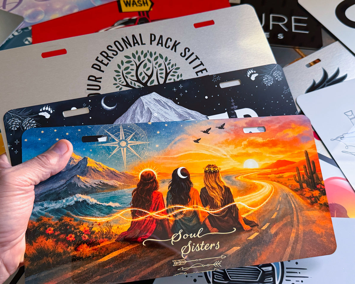 A hand holds a custom license plate featuring an artistic landscape of three women sitting by a desert road at sunset, with the text 'Soul Sisters' and '2025'. Other license plates are stacked in the background.