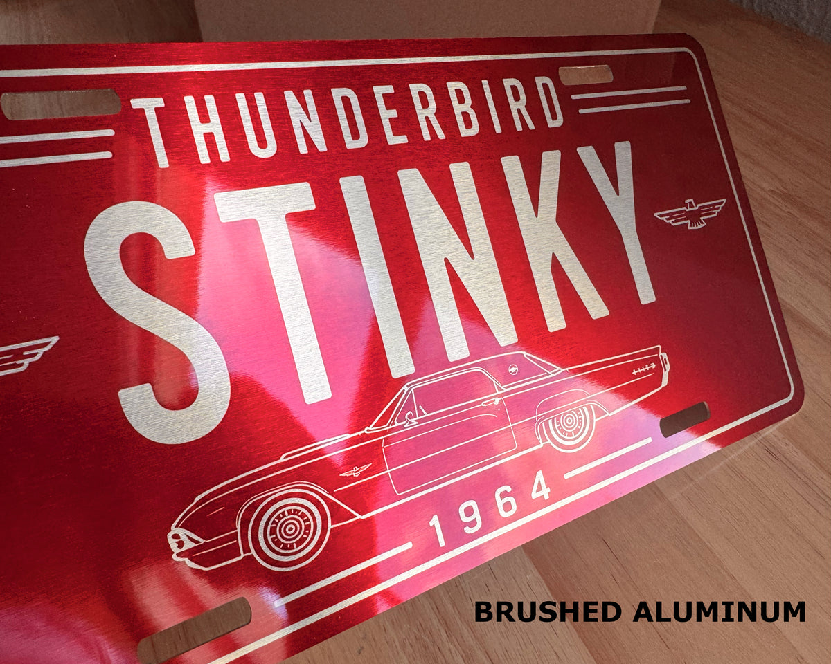 A red brushed aluminum novelty license plate featuring the words 'THUNDERBIRD STINKY 1964' and a white line drawing of a classic car, photographed on a wooden surface.