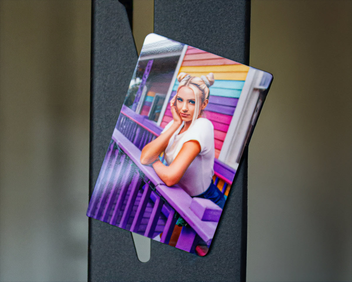 A rectangular printed  magnet with rounded corners, shown leaning against a dark vertical surface. The printed design features a stylized portrait of a blonde woman sitting on a purple porch in front of a rainbow-colored house.