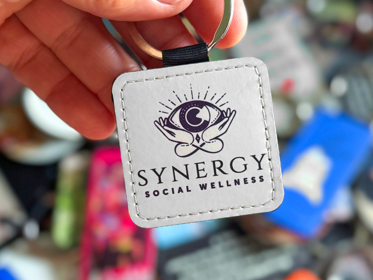 A close-up of a hand holding a square white leather keychain with a black stitched border and a black ribbon loop. The keychain features a black printed logo of hands holding an eye with rays, above the text 'SYNERGY SOCIAL WELLNESS'.