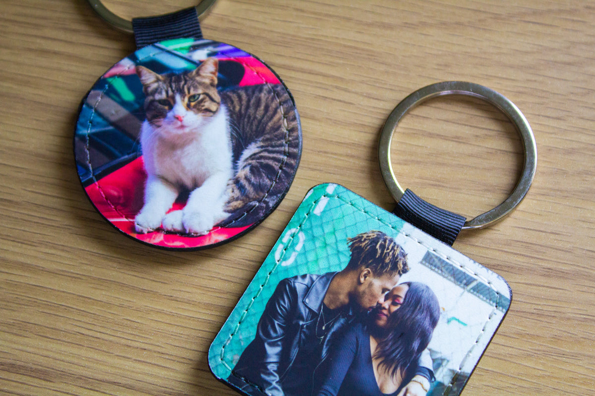 Two custom photo leather keychains on a light wood background. One round keychain features a photo of a tabby and white cat on a colorful background, and the other square keychain features a photo of a couple with a teal textured background.