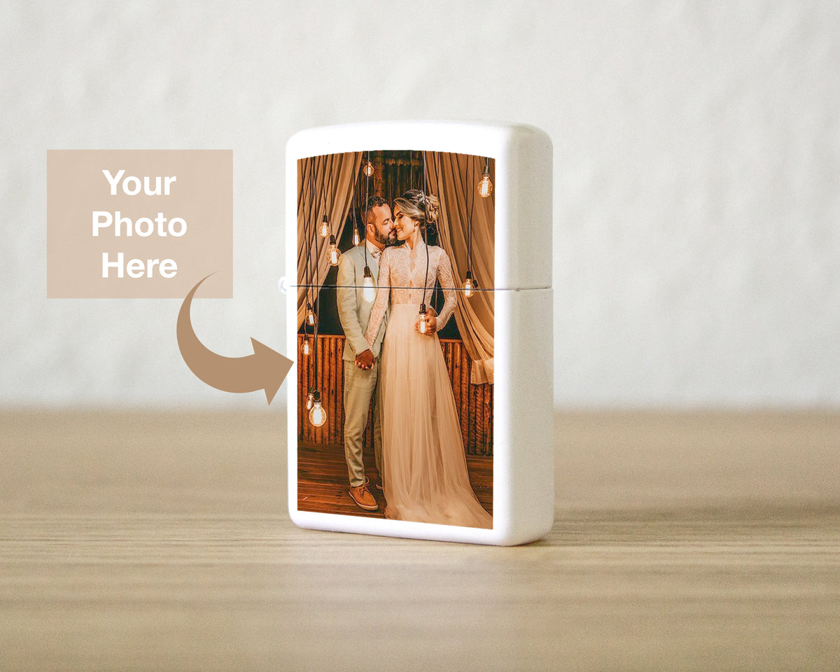 A white rectangular lighter or case standing on a light wood surface against a soft beige wall. The front has a printed custom photo of a couple embracing, with a tan placeholder box and arrow reading 'Your Photo Here' indicating customization.