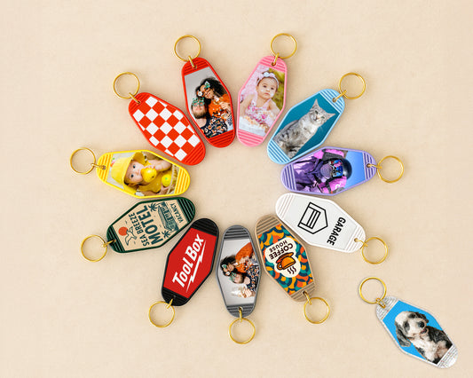 A set of colorful custom-shaped keychains arranged in a circular pattern on a light beige background. Each keychain features a different design, including novelty logos (such as Honda, Ford, and Harley-Davidson), brand names (Tim Tam, Hot Topic, D23), and pet photos.