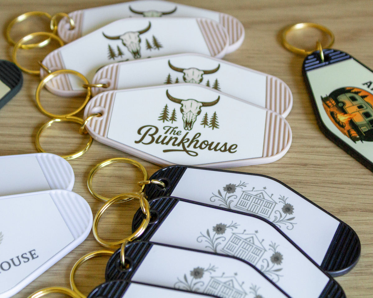 A close-up of several custom acrylic keychains with gold-tone keyrings on a light wood surface. The keychains feature rustic illustrations of a bison skull, pine trees, a house, and floral motifs, with some printed in white and others in black and white.