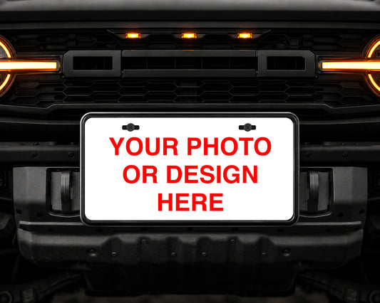 A customizable license plate mounted on a black vehicle grille, featuring a white background with red placeholder text that reads 'YOUR PHOTO OR DESIGN HERE'.