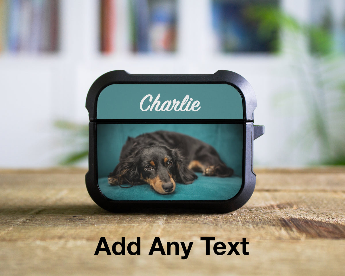 A custom protective AirPods Pro 1st 2nd or 3rd Gen case with a black bumper and a teal background, featuring a printed photo of a dachshund named 'Charlie' and the text 'Charlie' in white script. The case is shown on a wooden table with a blurred indoor background, and the text 'Add Any Text' is displayed below.