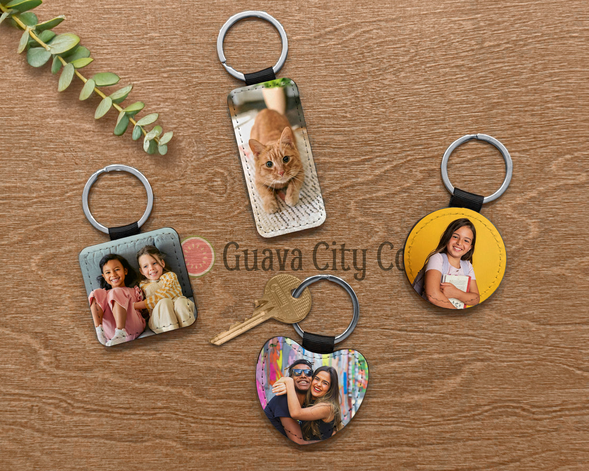 Four custom photo keychains displayed on a wooden surface, including square, round, and heart-shaped acrylic designs with printed photos, metal key rings, and a small set of keys. A green leafy sprig is in the top-left corner, and a watermark reading 'Guava City Co' is visible across the center.