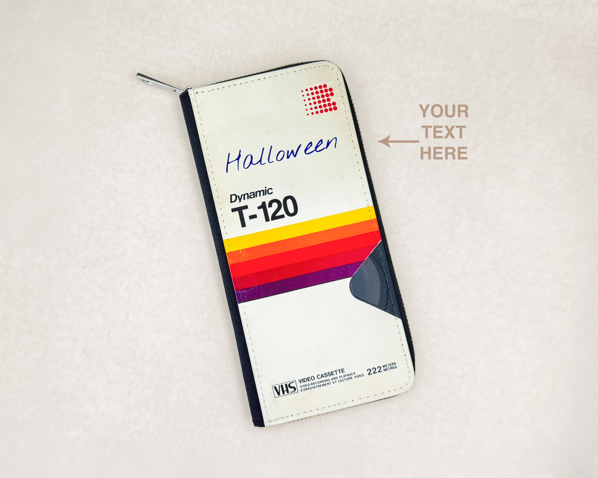 A custom zippered wallet styled as a VHS cassette sleeve T-120 , featuring a white background with a red grid logo, 'Halloween' written in blue script, and a retro rainbow stripe design. The wallet is shown on a textured light beige background with a text placeholder overlay.