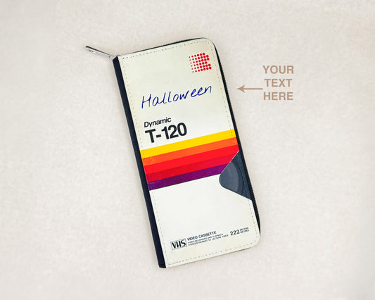 A custom zippered wallet styled as a VHS cassette sleeve T-120 , featuring a white background with a red grid logo, 'Halloween' written in blue script, and a retro rainbow stripe design. The wallet is shown on a textured light beige background with a text placeholder overlay.