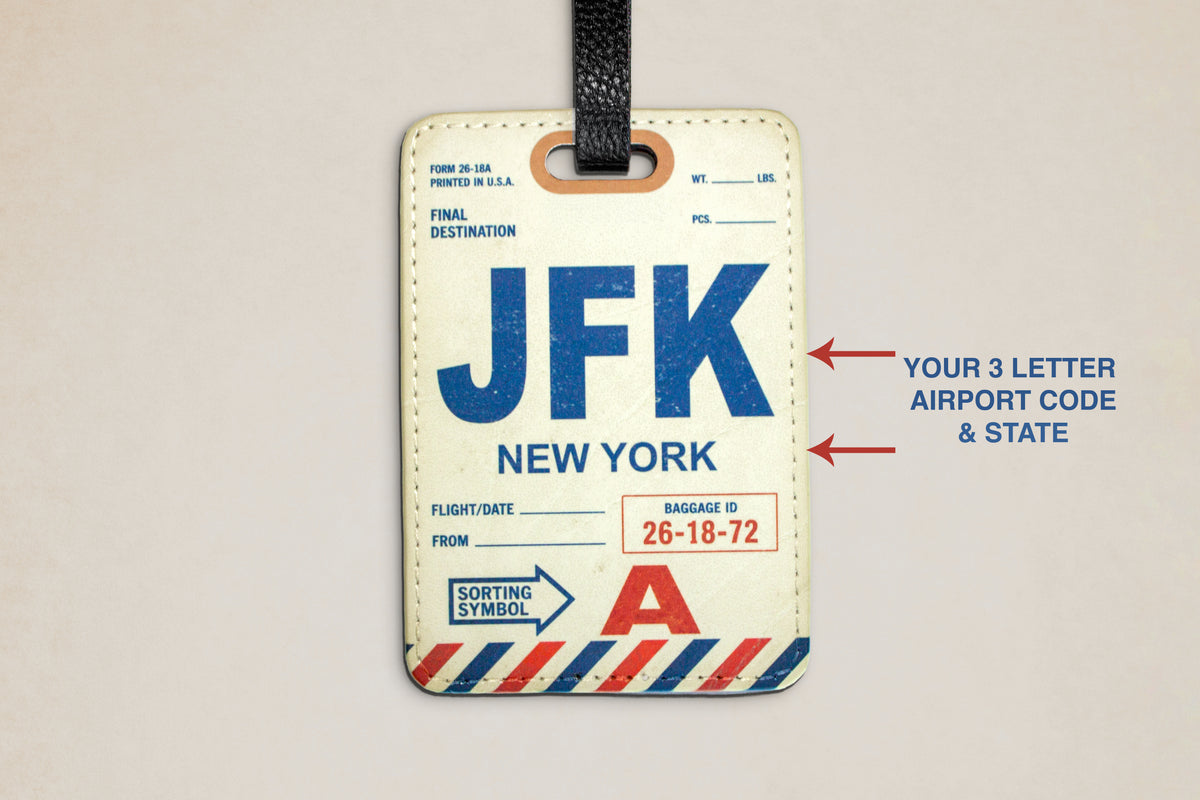 A cream-colored luggage tag with blue and red printed details, featuring the text 'JFK NEW YORK' and a black hanging strap. Red arrows on the right point to the 'JFK' airport code and state example.