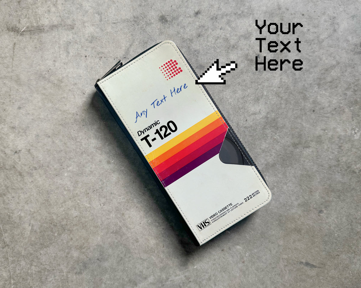 A custom printed wallet or card holder shown on a gray concrete floor, with a white faux-leather cover featuring a retro VCR or VHS cassette box orange, yellow, and red stripe graphic, placeholder text, An arrow graphic points to the cover with the text 'Your Text Here'. This is where customer can personalized the product with their own text.