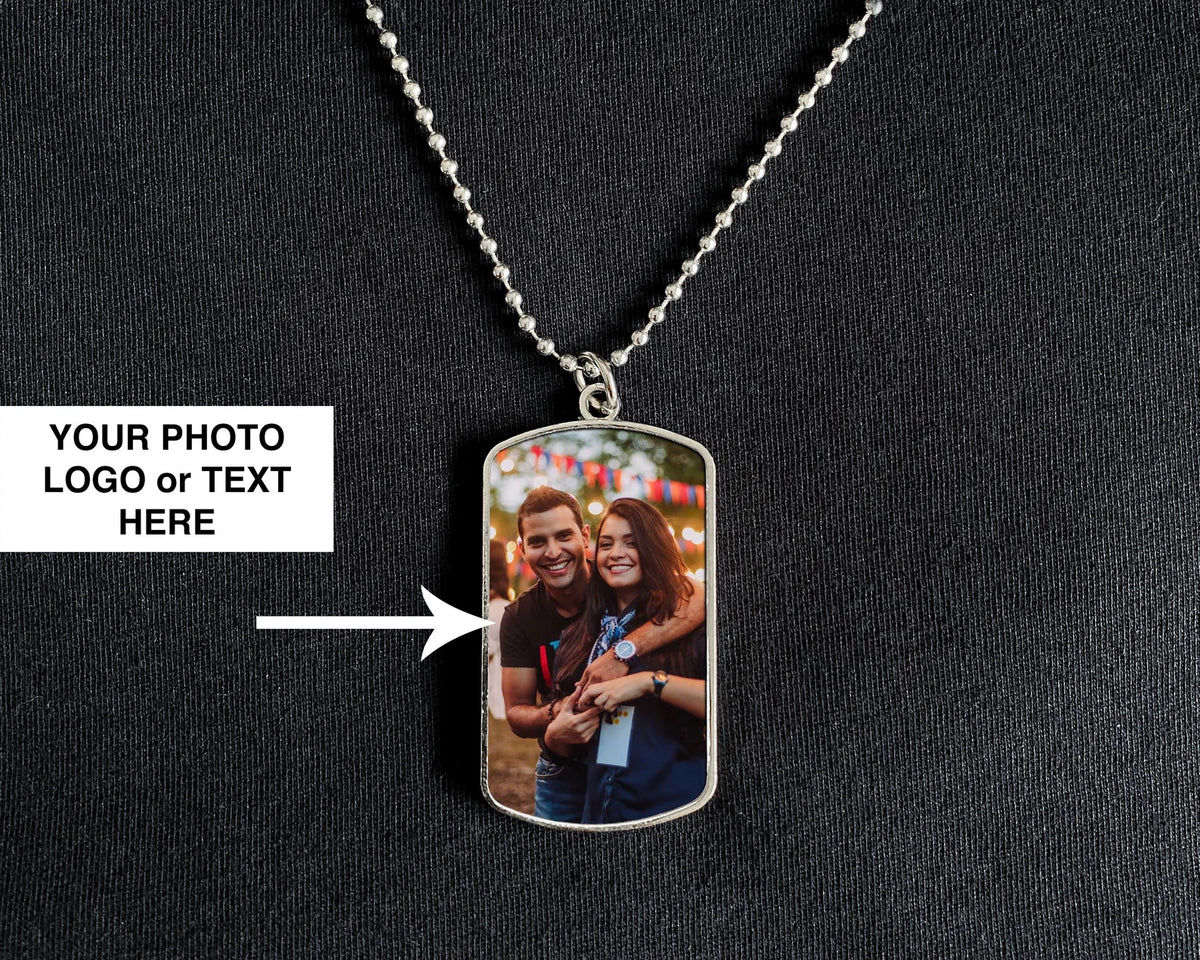 A silver dog tag pendant necklace displayed on a black textured fabric background. The pendant features a custom printed photo of a couple, with a white text box on the left reading 'YOUR PHOTO LOGO or TEXT HERE' and an arrow pointing to the photo area.