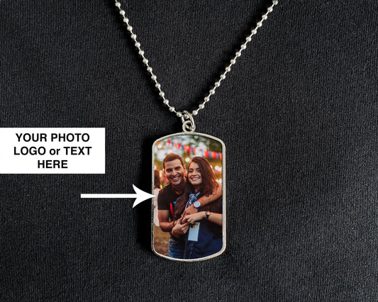 A silver dog tag pendant necklace displayed on a black textured fabric background. The pendant features a custom printed photo of a couple, with a white text box on the left reading 'YOUR PHOTO LOGO or TEXT HERE' and an arrow pointing to the photo area.